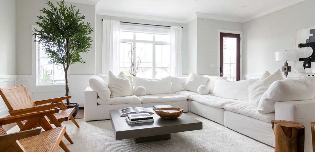 A bright living room with a large white sectional sofa, wooden chairs, and a ceiling fan and light providing soft illumination above.