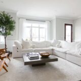 A bright living room with a large white sectional sofa, wooden chairs, and a ceiling fan and light providing soft illumination above.