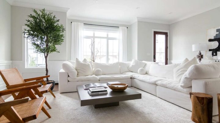 A bright living room with a large white sectional sofa, wooden chairs, and a ceiling fan and light providing soft illumination above.